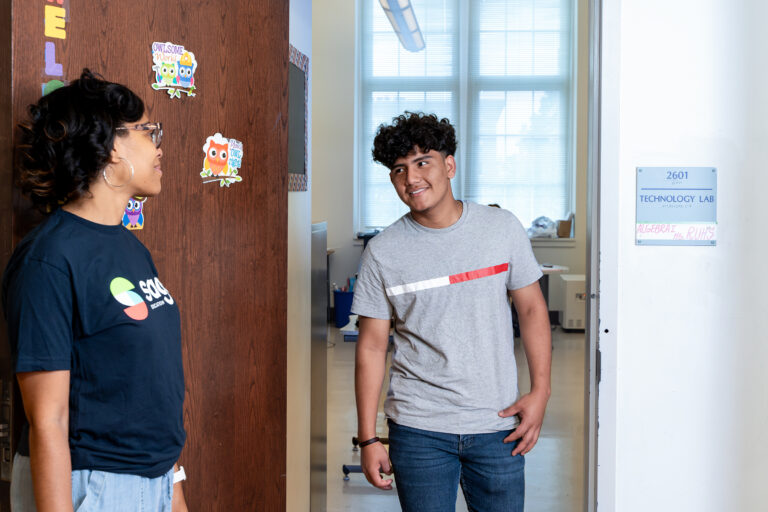 Two people, a tutor and a student, stand in front of a door. The tutor smiles at the student in the doorway of a classroom.